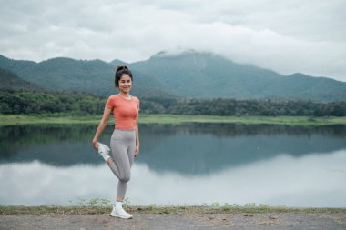 Aktif kıyafetli bir kadın, sakin bir gölün kenarında geriniyor, arkasında dağlar ve bulutlu bir gökyüzü var..