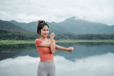 Aktif kıyafetli genç bir kadın bulutlu bir gökyüzünün altında arka planda dağlarla sakin bir göl kenarında esniyor..