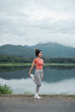 Aktif kıyafetli bir kadın gölün kenarında geriniyor. Arka planda dağlar var. Spor salonu ve doğa..