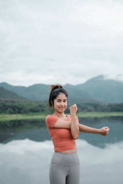 Aktif kıyafetli bir kadın bulutlu bir gökyüzünün altında arka planda dağlarla sakin bir göl kenarında geriniyor..