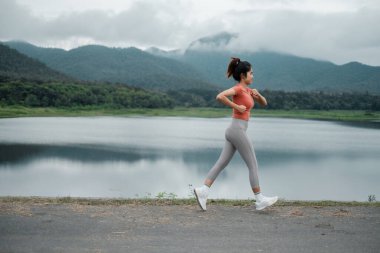 Arka planda dağlar olan göl kenarında koşan bir kadın, fitness ve doğayı destekliyor..