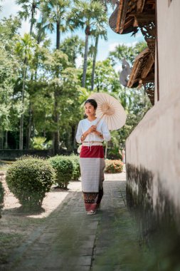 Geleneksel giyinmiş bir kadın, yemyeşil ve tarihi mimariyle çevrili canlı bir bahçede şemsiyeyle yürüyor..