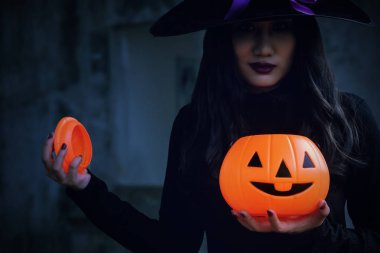 Genç kadın cadı Halloween kılık korkutucu koyu arka plan üzerinde turuncu kabak tutun.