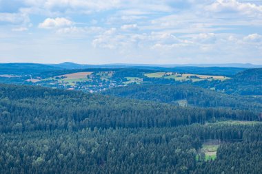 Yoğun ormanlarla kaplı dağlık alan, arka planda küçük bir köy ve buğday tarlaları, sabahın erken saatlerinde.