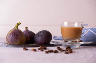 Sütlü bir fincan aromatik kahve ve 3 olgun incir, kahve çekirdekleri dağılmış ve bunların hepsi desenli gri bir arka planda.