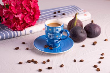 Mavi kahve fincanı, dağılmış kahve çekirdekleri, arka planda 3 incir ve ortanca dalı, desenli gri arka plan.