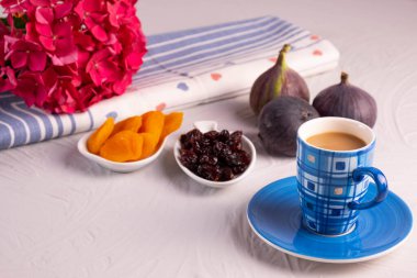 Sütlü bir fincan kahve, kurutulmuş kayısı ve kızılcık, taze incir, bir peçetedeki pembe ortanca dalı, bunların hepsi dokulu arka planda, gri