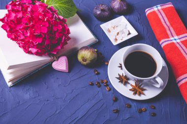 Bir fincan kahve, kahve çekirdekleri ve şeker küpleri, açık bir kitap ve bir dal ortanca, mavi arka planda üç incir.