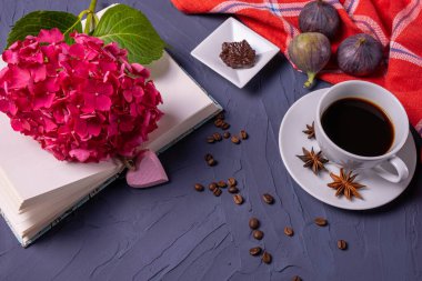 aromaton kahve, açık kitap ve pembe ortancalar, kırmızı peçetede üç incir, dağınık kahve çekirdekleri ve mavi arka planda koyu çikolata parçaları.