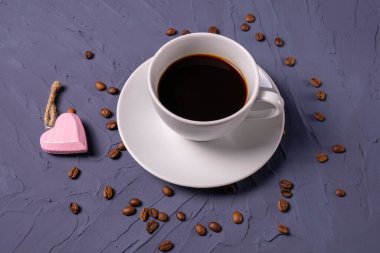 Bir fincan sıcak ve aromatik kahve serpiştirilmiş kahve çekirdekleri ve dokulu mavi arka planda kalp şeklinde kolye.