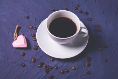 Bir fincan sıcak ve aromatik kahve, kahve çekirdeği ve dokulu mavi arka planda kalp şeklinde bir kolye.