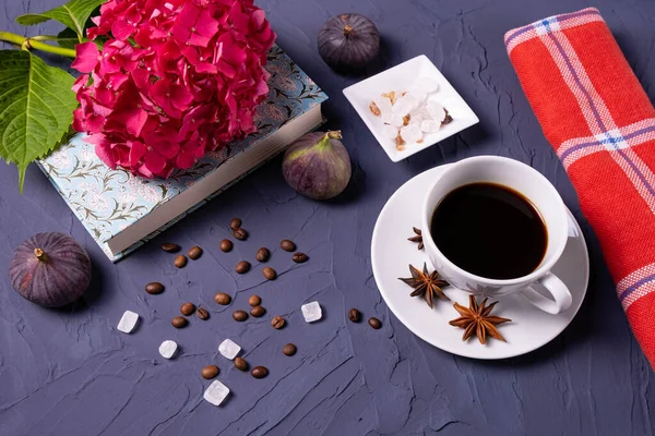 Bir fincan kahve, kahve çekirdekleri ve şeker küpleri, bir kitap ve bir dal ortanca, mavi arka planda üç incir.