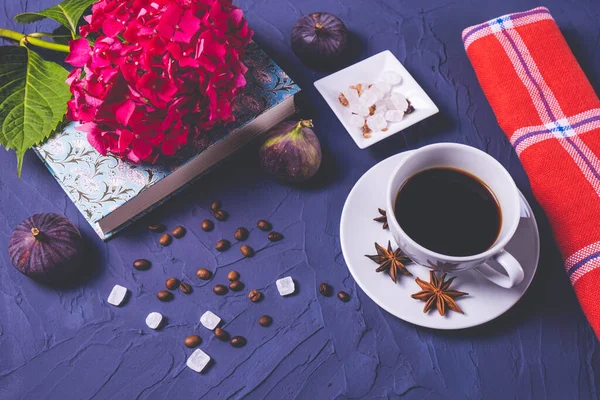 Bir fincan kahve, dağınık kahve çekirdekleri ve şeker, üç incir, anason, kapalı bir kitap ve üzerine bir dal ortanca.