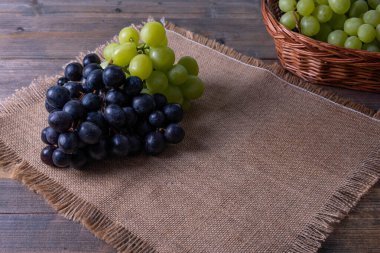Hasat edilmiş olgun üzümler bir sepetin içinde ahşap bir masanın üzerinde ve üzüm demetleri keten bir masa örtüsünün üzerinde yatıyor.