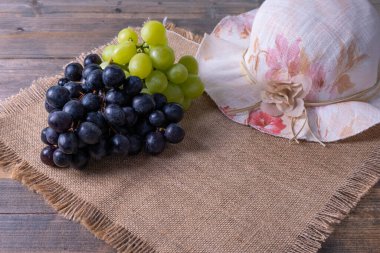 Olgun sulu üzüm hasadı demetleri, kadın şapkası ahşap bir masada ve pürüzlü bir masa örtüsünde yatıyor.