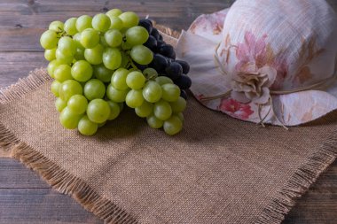 Olgun, sulu, yeşil ve siyah üzüm ve bir kadının şapkası ahşap bir masanın üzerinde duruyor.