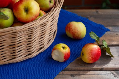 Mavi masa örtüsünün üzerinde üç tane elma var. Bir tanesi çoktan ısırılmış ve yanında bir sepet dolusu elma var.