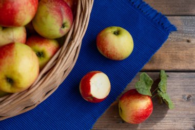 Üç elma, biri çoktan ısırılmış, mavi bir masa örtüsünün üzerinde ve dolu bir sepet elmanın yanında yatıyor.