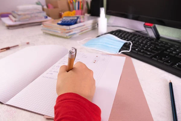 at the desk, hand holds a pen and writes in a notebook in a cage, mathematical solutions, textbooks and a mask with disinfectant in the background