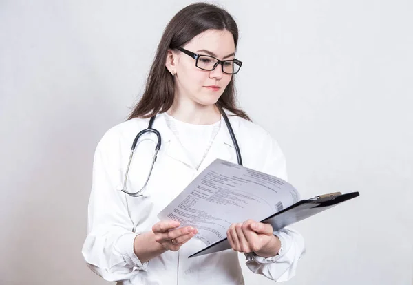 Retrato de uma jovem mulher sorridente médico morena em óculos e um casaco branco médico com um estetoscópio e uma pasta para papéis em um fundo branco — Fotografia de Stock