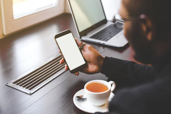 Hombre afroamericano sentado en un café, bebiendo café y trabajando detrás de un teléfono móvil. Mock up pantalla — Foto de Stock