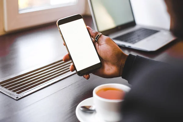 Hombre afroamericano sentado en un café, bebiendo café y trabajando detrás de un teléfono móvil. Mock up pantalla — Foto de Stock