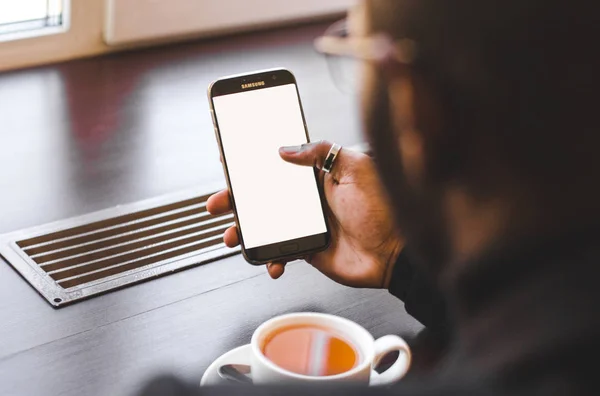 Hombre afroamericano sentado en un café, bebiendo café y trabajando detrás de un teléfono móvil. Mock up pantalla — Foto de Stock