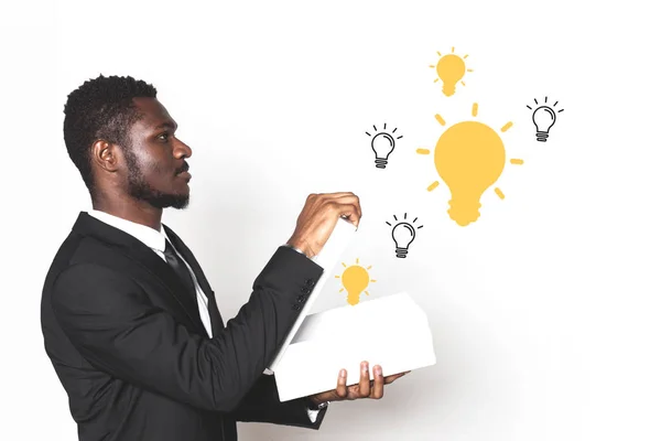 Empresario afroamericano en un traje sostiene una caja de la que las ideas vienen en forma de bombillas. aislado sobre fondo blanco —  Fotos de Stock