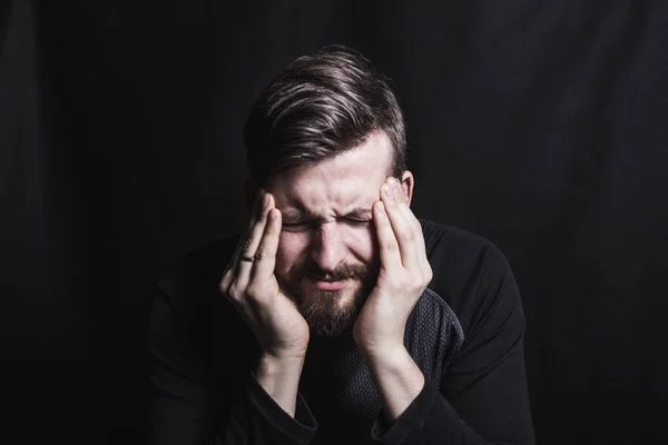 Um homem segura a cabeça e sofre de uma dor de cabeça, esfrega suas têmporas com os dedos em um fundo escuro — Fotografia de Stock