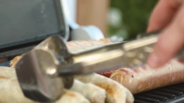Barbacoa con salchichas ardientes en la parrilla en un picnic al aire libre. primer plano de las manos femeninas con pinzas — Vídeos de Stock