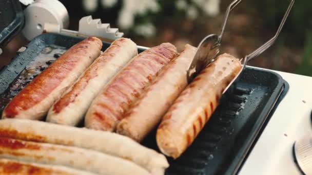 Churrasco com salsichas de fogo na grelha em um piquenique ao ar livre. close-up de mãos femininas com pinças — Vídeo de Stock