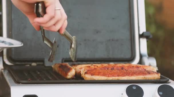 BBQ dengan sosis berapi-api di panggangan pada piknik di luar ruangan. close-up dari tangan perempuan dengan penjepit — Stok Video