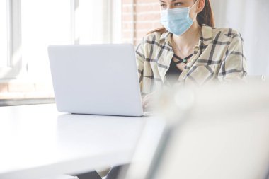 Coronavirus covid-19 için tıbbi maskeli beyaz bir kadın laptopla ofiste çalışıyor.