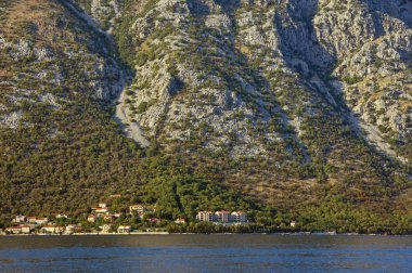 Eteğindeki küçük sahil köyü ile güneş tarafından aydınlatılmış sarp dağ yamacı doku. Karadağ, Kotor Körfezi