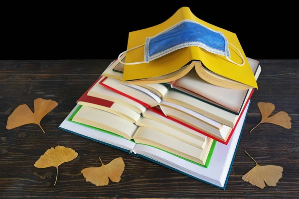 Open books, yellow leaves and face protective mask on the table. Black background, copy space