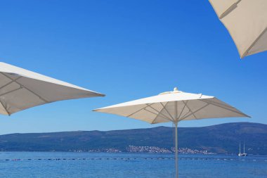 Plaj tatili kavramı. Mavi gökyüzüne karşı beyaz güneş şemsiyeleri. Karadağ, Adriyatik Denizi. Tivat kenti yakınlarındaki Kotor Körfezi manzarası
