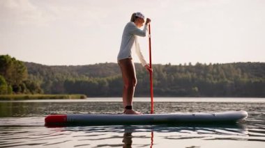 Bir insan kürek çekiyor, sakin sularda ustaca manevra yapıyor, doğal çevreyi yansıtıyor, macera duygusunu ve doğayla bağlantıyı somutlaştırıyor.