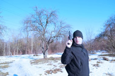 Siyah elbiseli genç bir adam, elinde silahla kışın doğasında.