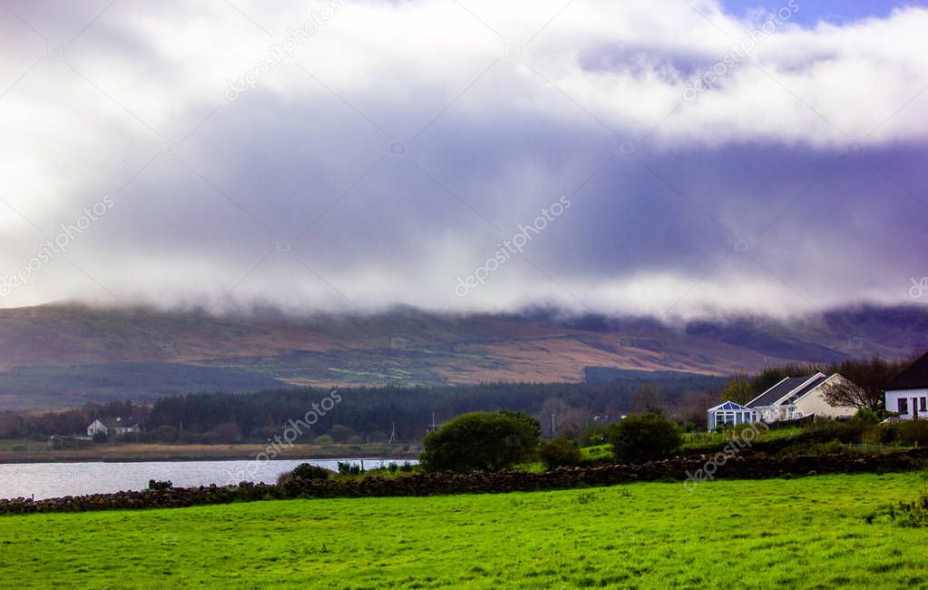 view of the sea shore in Ireland rain clouds hanging over the mountains and blue sky, sun showing from the cloud green fields river