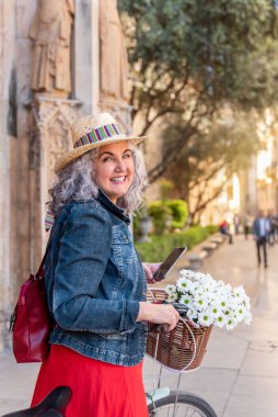 Gülümseyen yaşlı kadın emekliliğin tadını çıkarıyor. Bisiklete binerken sepetinde çiçekler vardı.