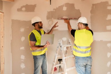 İki erkek inşaat işçisi bir evin yenilenmesi için dizüstü bilgisayarla çalışırken duvarı yıkıyorlar.