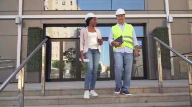 İnşaat alanındaki vinç yakınındaki planları inceleyen iki farklı sert şapka ve yelekli profesyonel.