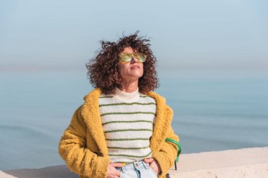 Gözleri kapalı İspanyol kadın, kıvırcık saçlı ve güneş gözlüklü deniz kenarında güneşlenen.