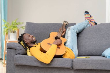 Gülümseyen Afrikalı Amerikalı adam evde bir kanepede yatarken gitar akoru çalışıyor.