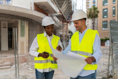 Güvenlik yelekleri ve inşaat alanındaki planı gözden geçiren iki farklı mimar.