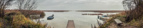 panoramik görünümü göle koyu ahşap bir iskele, ufukta sazlık ile bulutlu havalarda sonbahar alacakaranlık tekneler ile kıyı bitki örtüsü ile