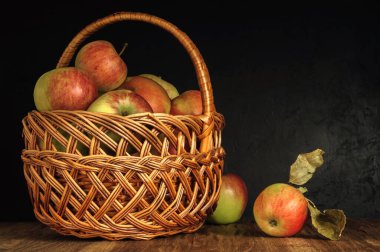 Kırmızı-yeşil olgun elmalar hasır bir sepetin içinde eski bir tahta masada siyah alçıdan yapılmış bir arka planda. Yan görünüş. Sonbahar kavramsal karanlık fotokopi alanı ile kırsal tarzda yaşam