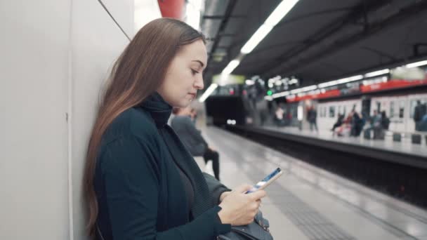 Жінка друкує на смартфоні на станції метро. Очікування на поїзд — стокове відео