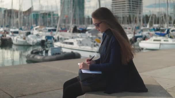 Mujer dibujando bocetos en su cuaderno al aire libre. Palmeras y yates sobre fondo — Vídeo de stock