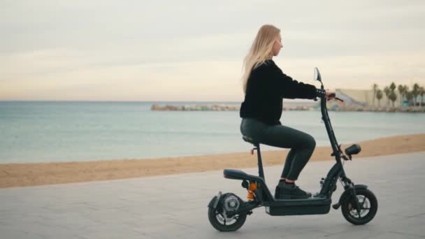 Mujer rubia bonita conduciendo bicicleta eléctrica cerca del mar . — Vídeos de Stock